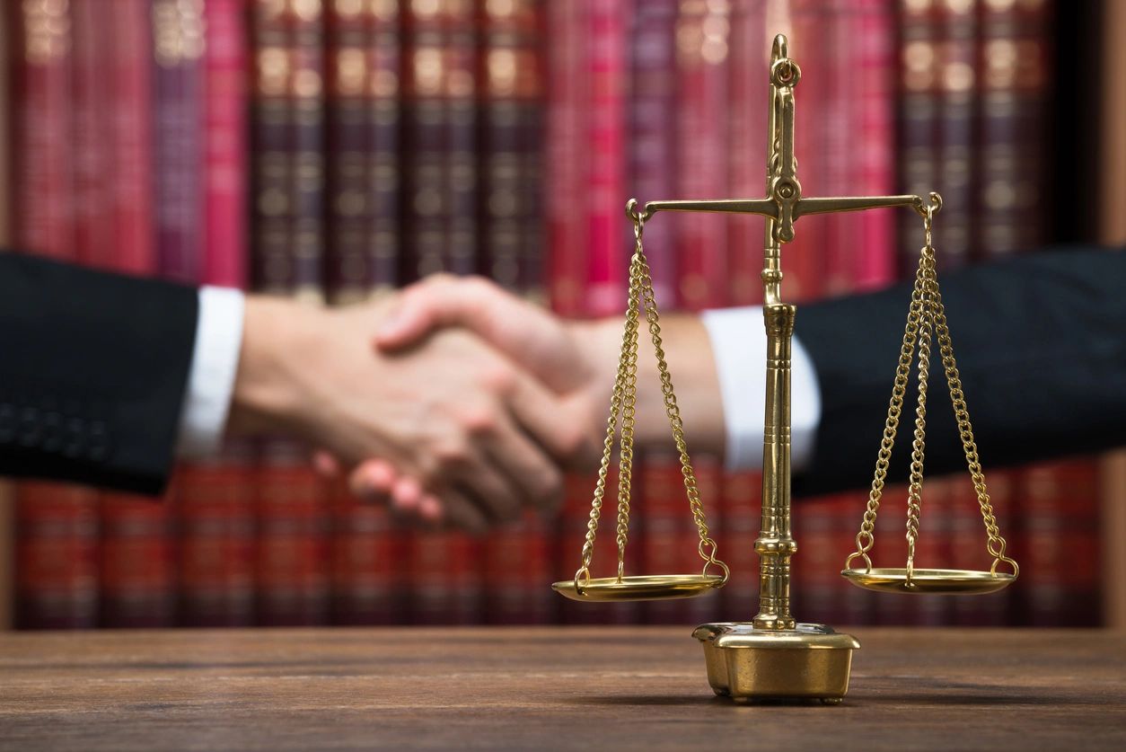 Justice Scale on Wooden Table With Legal Adviser and Client Shaking Hands in Background at Courtroom